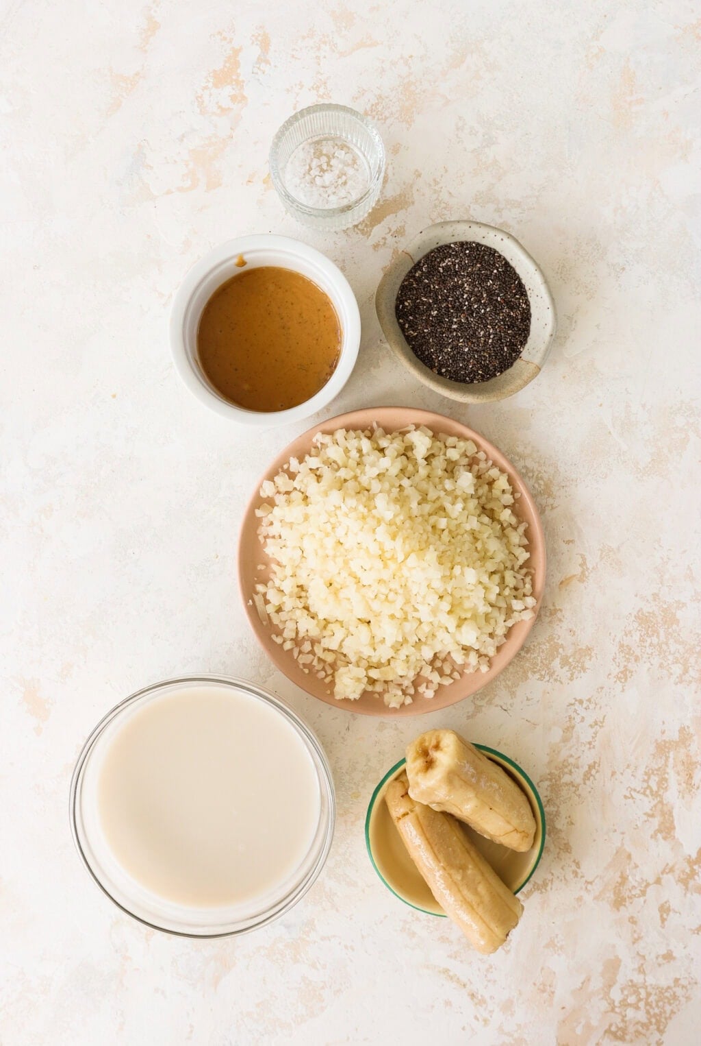 Six bowls containing various ingredients are arranged on a light surface. The bowls hold salt, chia seeds, nut butter, cauliflower rice, banana pieces, and a white liquid (likely plant-based milk).