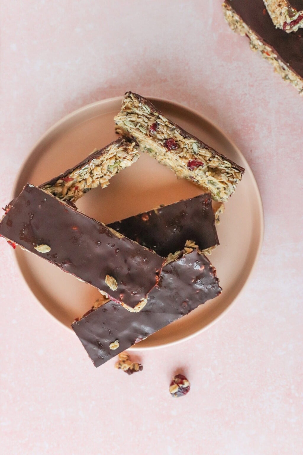 Five chocolate-covered granola bars with nuts and dried fruit are arranged on a round plate against a pink background. Some nuts and dried fruit pieces are scattered nearby.