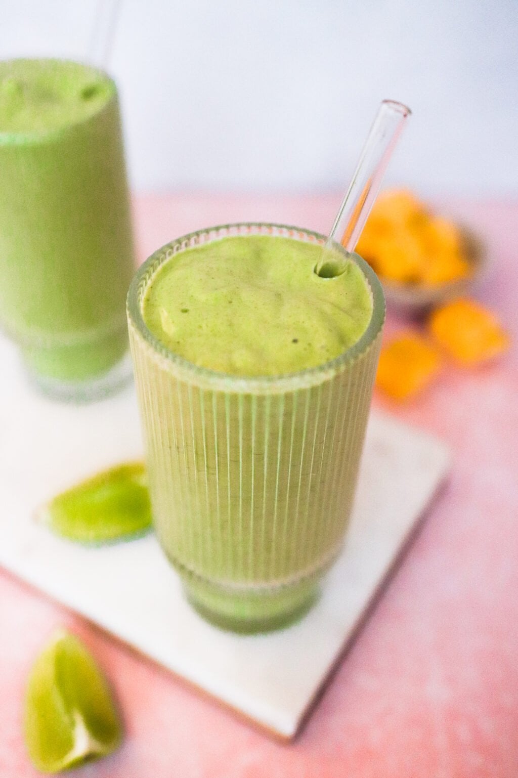 A glass of green smoothie with a clear straw sits on a white board, with lime wedges and a bowl of orange fruit pieces in the background. Another glass of green smoothie is partially visible beside it.