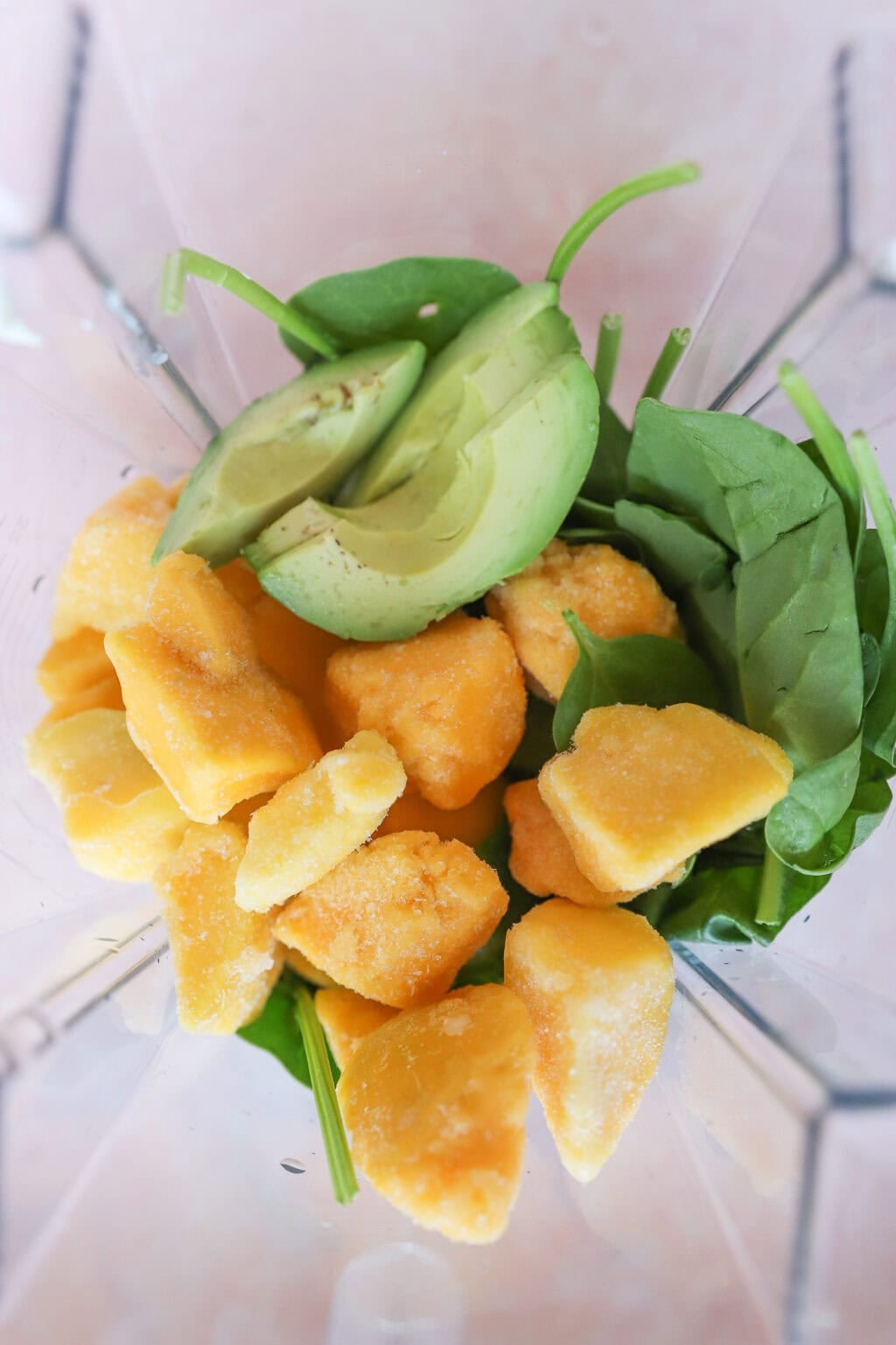 A top-down view of a blender containing fresh spinach leaves, sliced avocado, and chunks of frozen mango, ready to be blended.