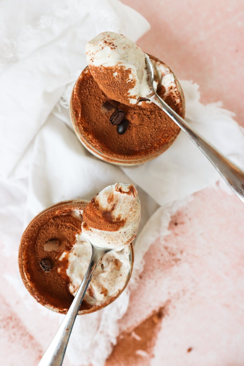 Two metal spoons holding creamy tiramisu are placed above two small ramekins filled with tiramisu topped with cocoa powder and coffee beans, set on a white cloth and a pink surface.