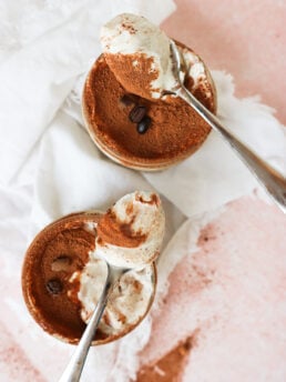 Two metal spoons holding creamy tiramisu are placed above two small ramekins filled with tiramisu topped with cocoa powder and coffee beans, set on a white cloth and a pink surface.