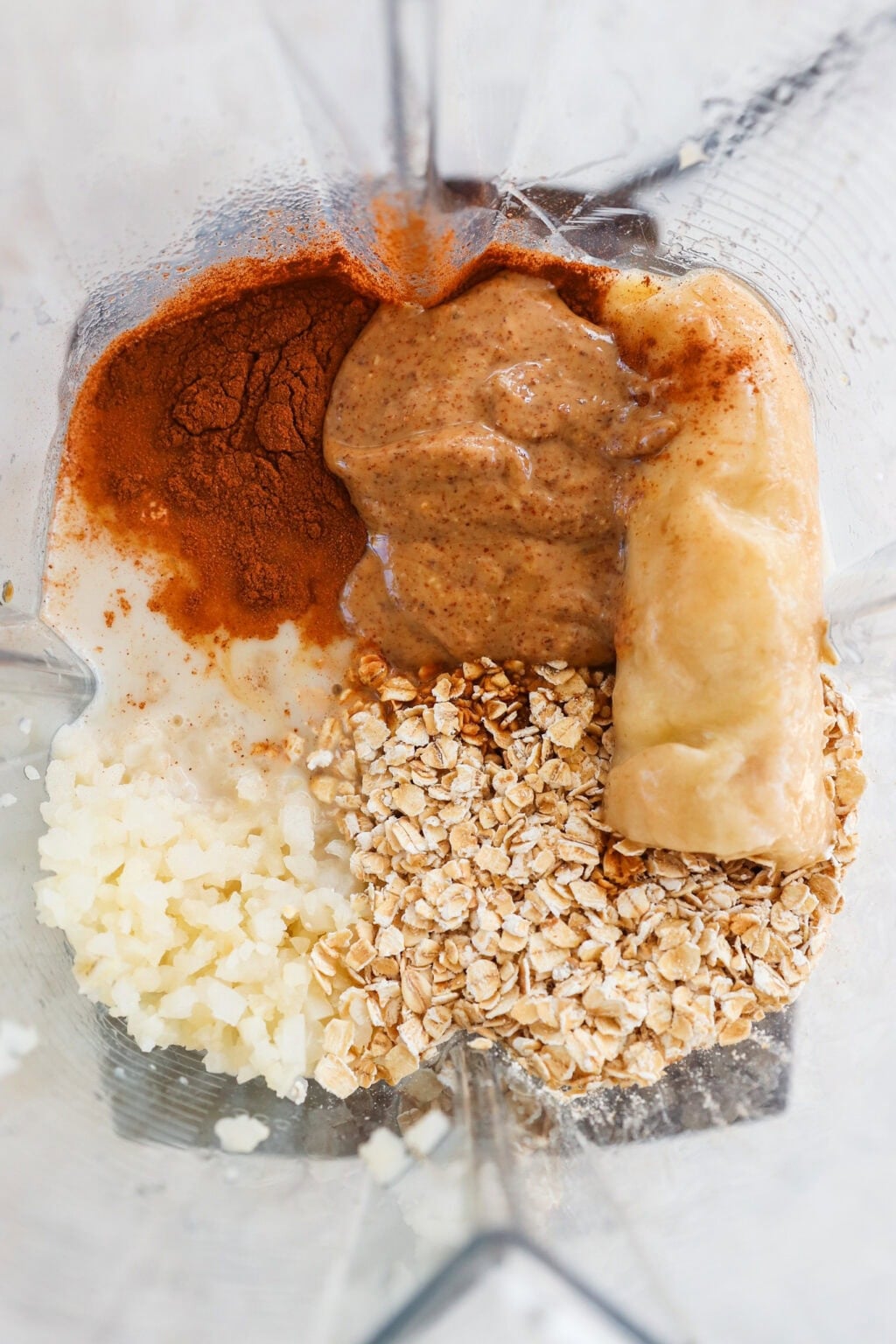 A close-up of a blender containing almond butter, a banana, oats, cauliflower rice, cinnamon, and a small amount of liquid, ready to be blended.
