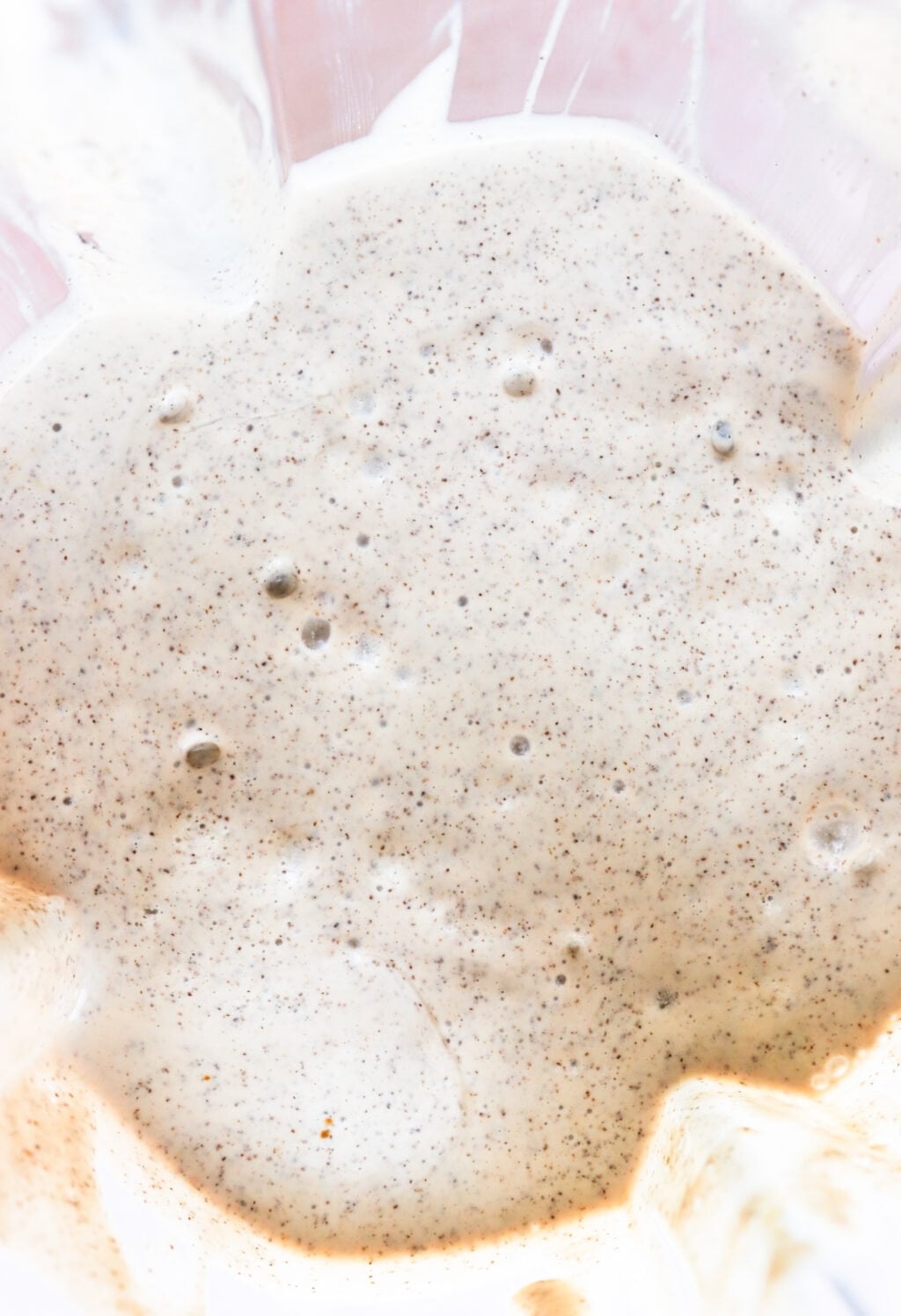 Close-up of bubbly, creamy batter with visible specks throughout, likely from whole grain or spices, in a mixing bowl. The mixture appears thick and airy.