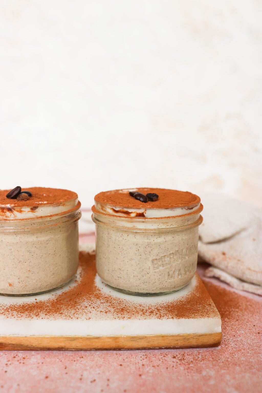 Two small glass jars filled with creamy tiramisu, topped with cocoa powder and a coffee bean, sit on a white wooden board. The background is softly blurred in beige and pink tones.