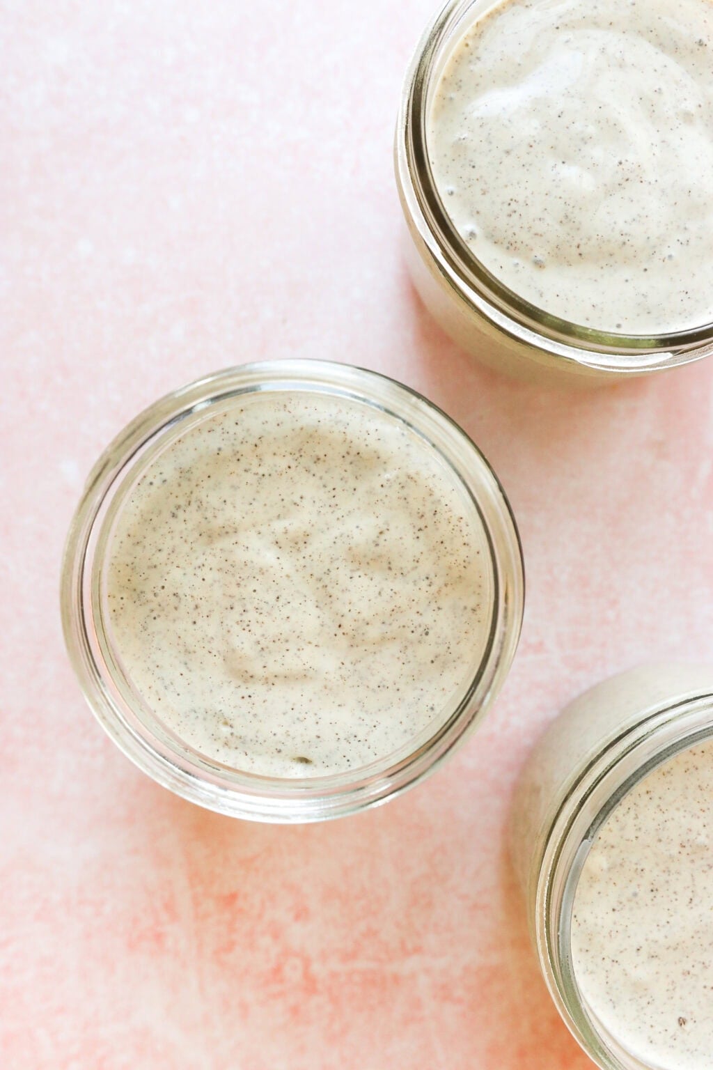 Three glass jars filled with a creamy, pale mixture speckled with tiny black vanilla bean flecks are arranged on a soft pink surface, viewed from above.