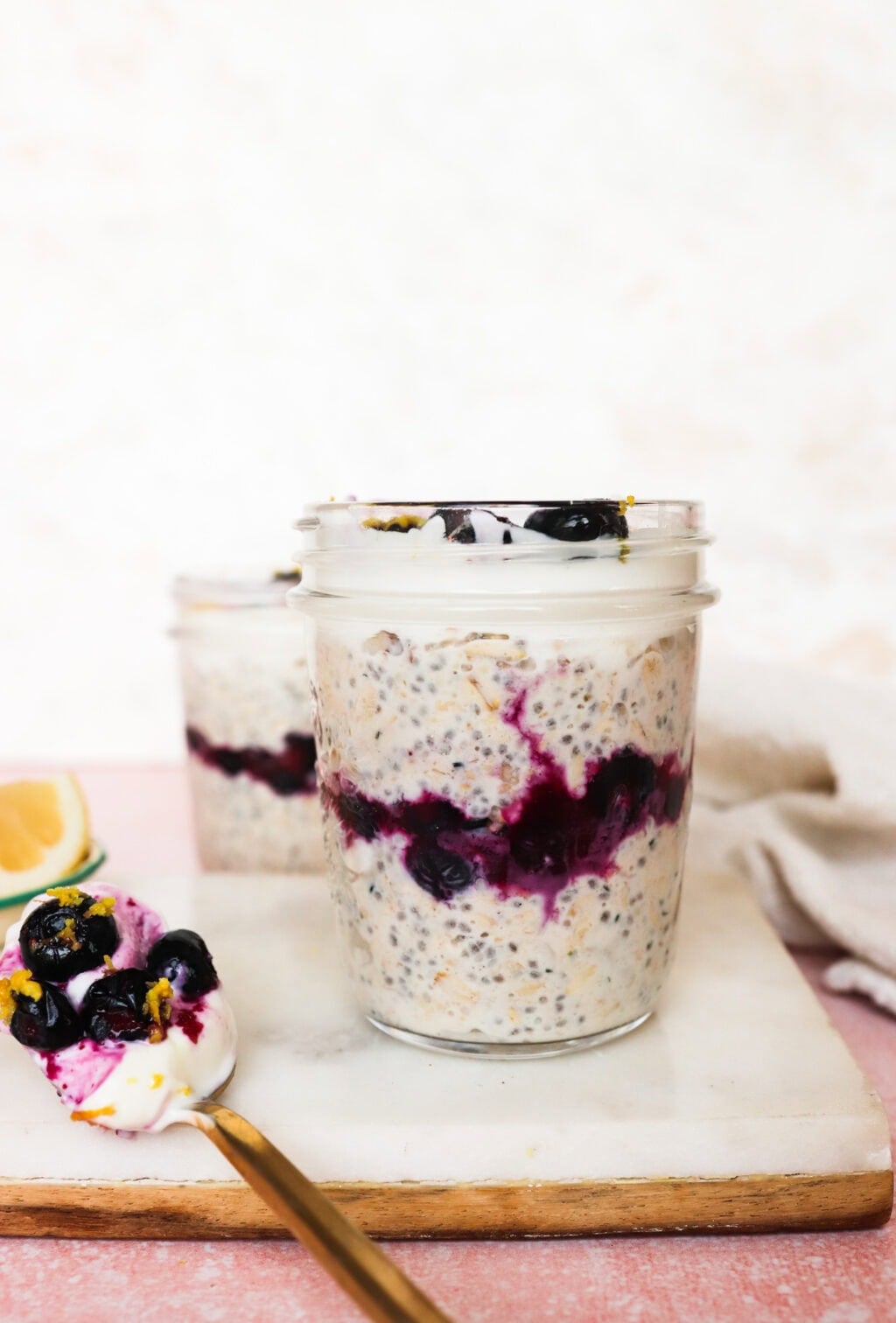 A glass jar filled with overnight oats, chia seeds, and a layer of blueberry sauce, topped with more blueberries. A spoon with oats and blueberries rests beside the jar on a marble board. Another jar is blurred in the background.