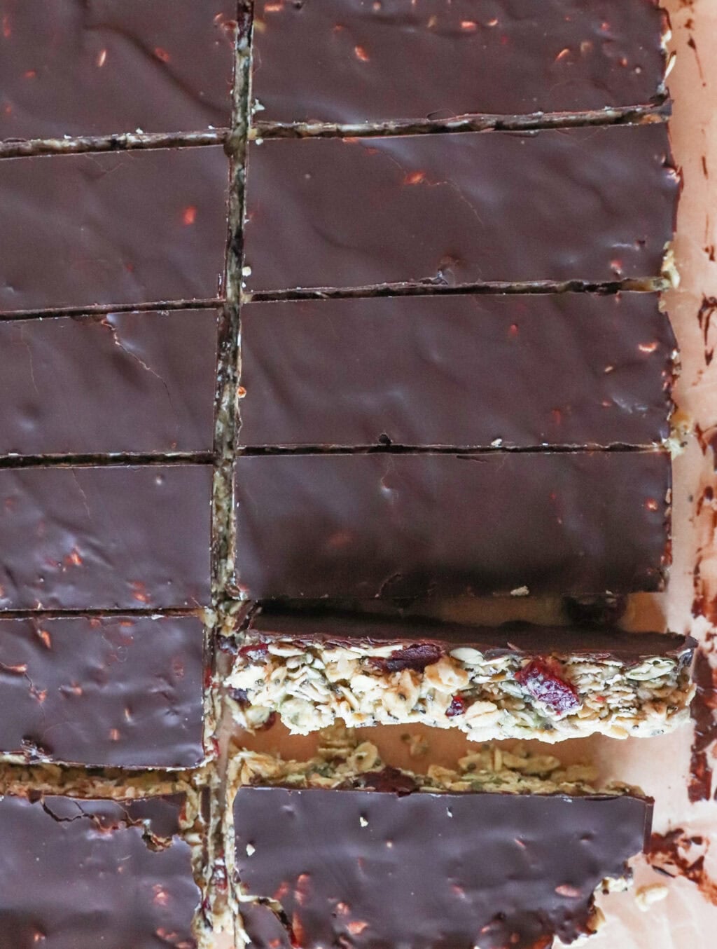 Close-up of chocolate-covered granola bars with visible nuts and oats on a cutting board. The bars are cut into rectangles, showing a rich, dark chocolate topping. One piece appears slightly pulled away from the rest.