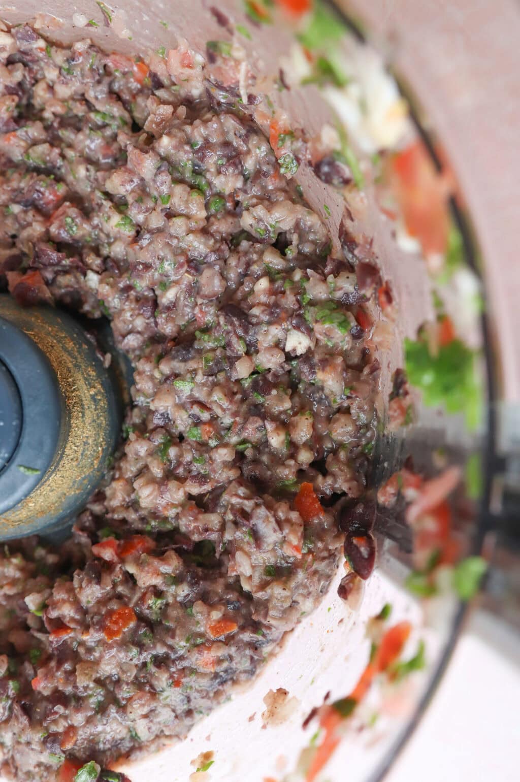Close-up of a food processor filled with a colorful mixture of chopped ingredients, including black beans, cilantro, tomatoes, and onions, creating a chunky texture. The image shows the circular motion of the blended ingredients.