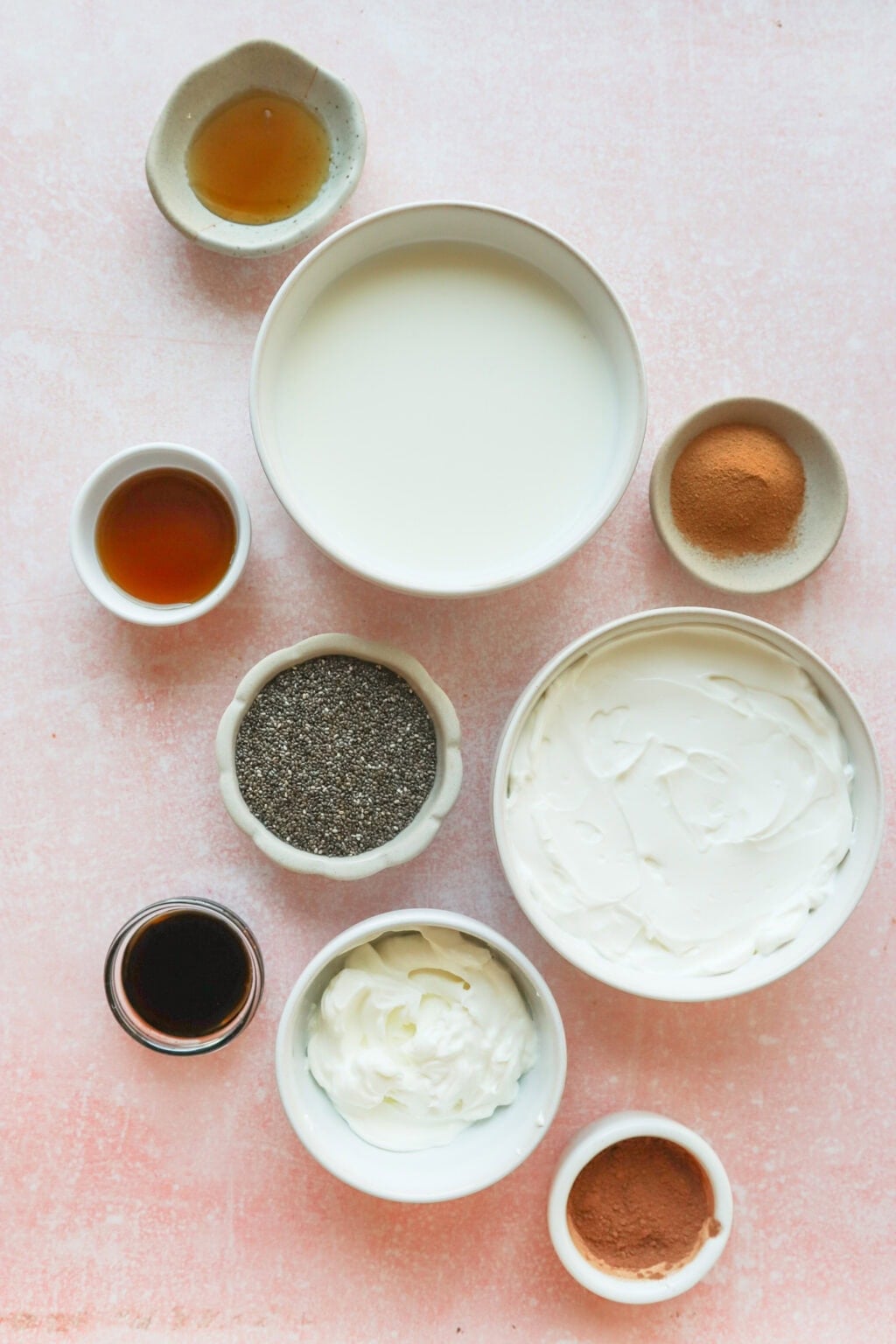 A flat lay of various ingredients in small bowls on a pink surface, including milk, yogurt, chia seeds, cocoa powder, greek yogurt, vanilla extract, and honey or syrup.