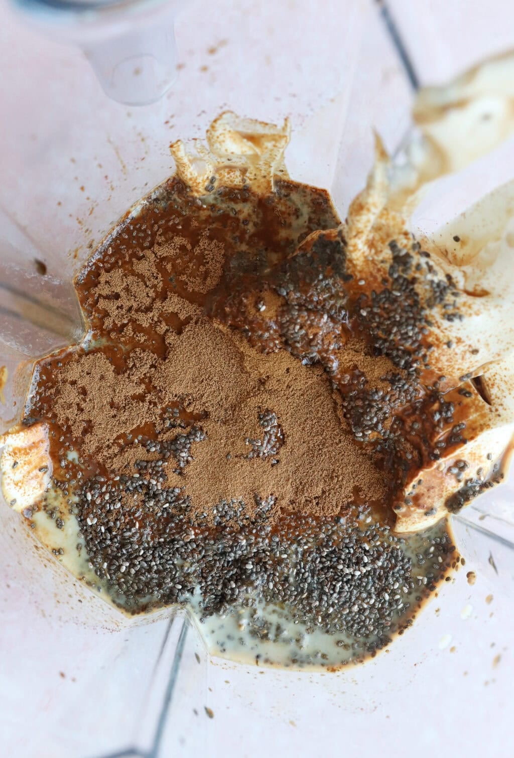 A close-up view from above of a blender containing chia seeds, cinnamon, a creamy liquid, and possibly protein powder, before blending.