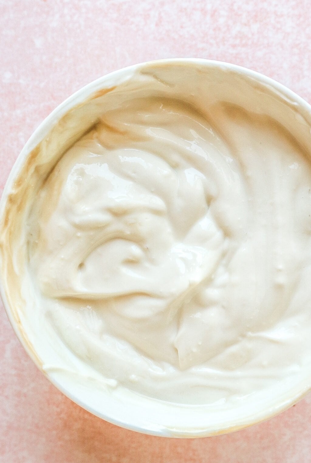 A close-up of a white bowl filled with creamy, smooth yogurt or a similar thick, white dairy product, placed on a light pink surface.