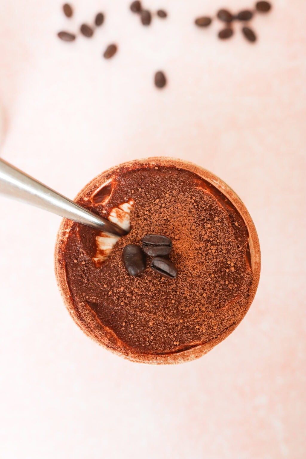 A top view of a chocolate dessert or drink topped with cocoa powder and coffee beans, with a spoon inside and scattered coffee beans in the background on a light pink surface.
