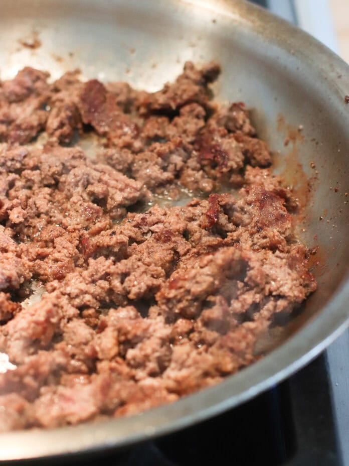 Ground beef is being cooked and browned in a stainless steel skillet on a stovetop. The meat is crumbled and sizzling, with some areas showing darker, crispy edges.