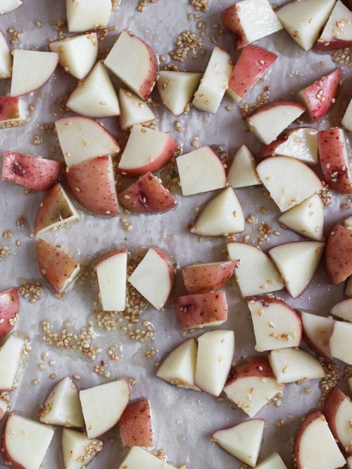 Chopped red potatoes with skins on, scattered on a parchment-lined baking sheet and sprinkled with sesame seeds, ready for roasting.