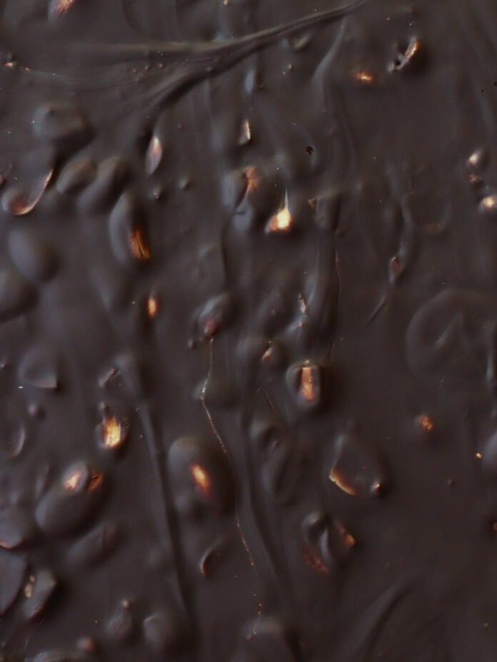 Close-up of a dark chocolate bar with visible nuts or crisped rice pieces embedded, creating a bumpy and textured surface with swirls and glossy areas.
