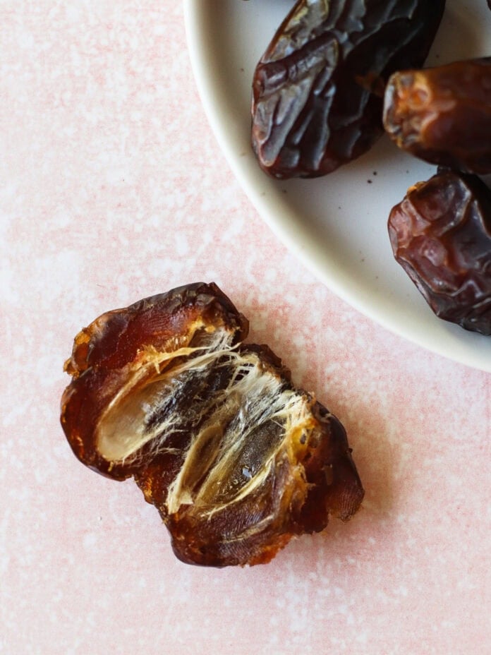 A close-up of a split open date fruit showing its fibrous interior and pit, next to a white plate with whole dates on a light pink textured surface.