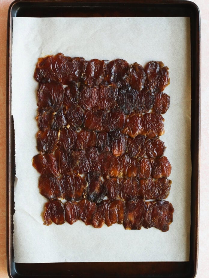 A rectangular layer of homemade fruit leather on parchment paper, placed on a dark baking tray. The fruit leather appears shiny, dark brown, and slightly wrinkled.