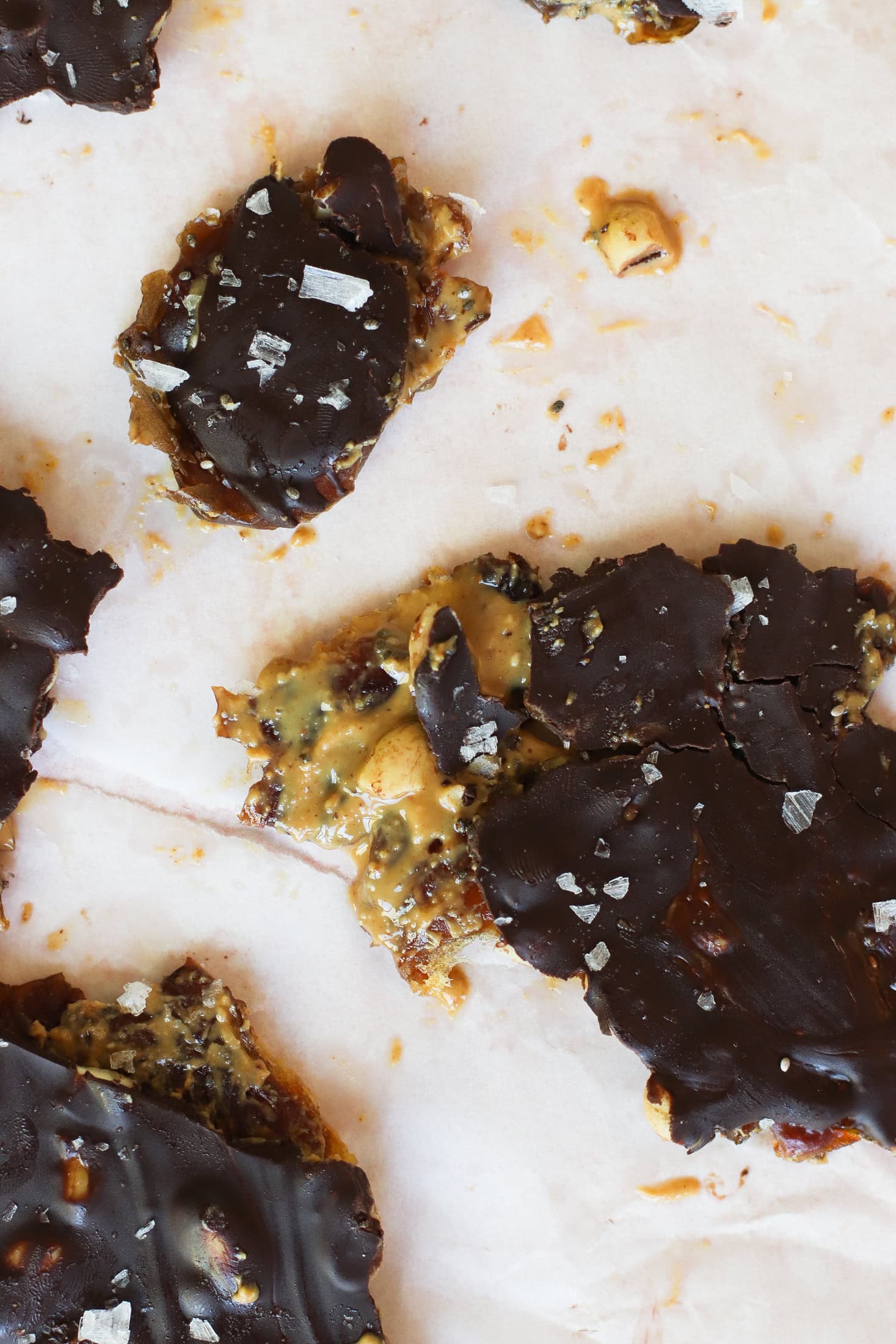 Close-up of pieces of chocolate bark with visible nuts and seeds, topped with flaky sea salt, on parchment paper. Some pieces are broken, revealing a caramel-like layer underneath the chocolate.
