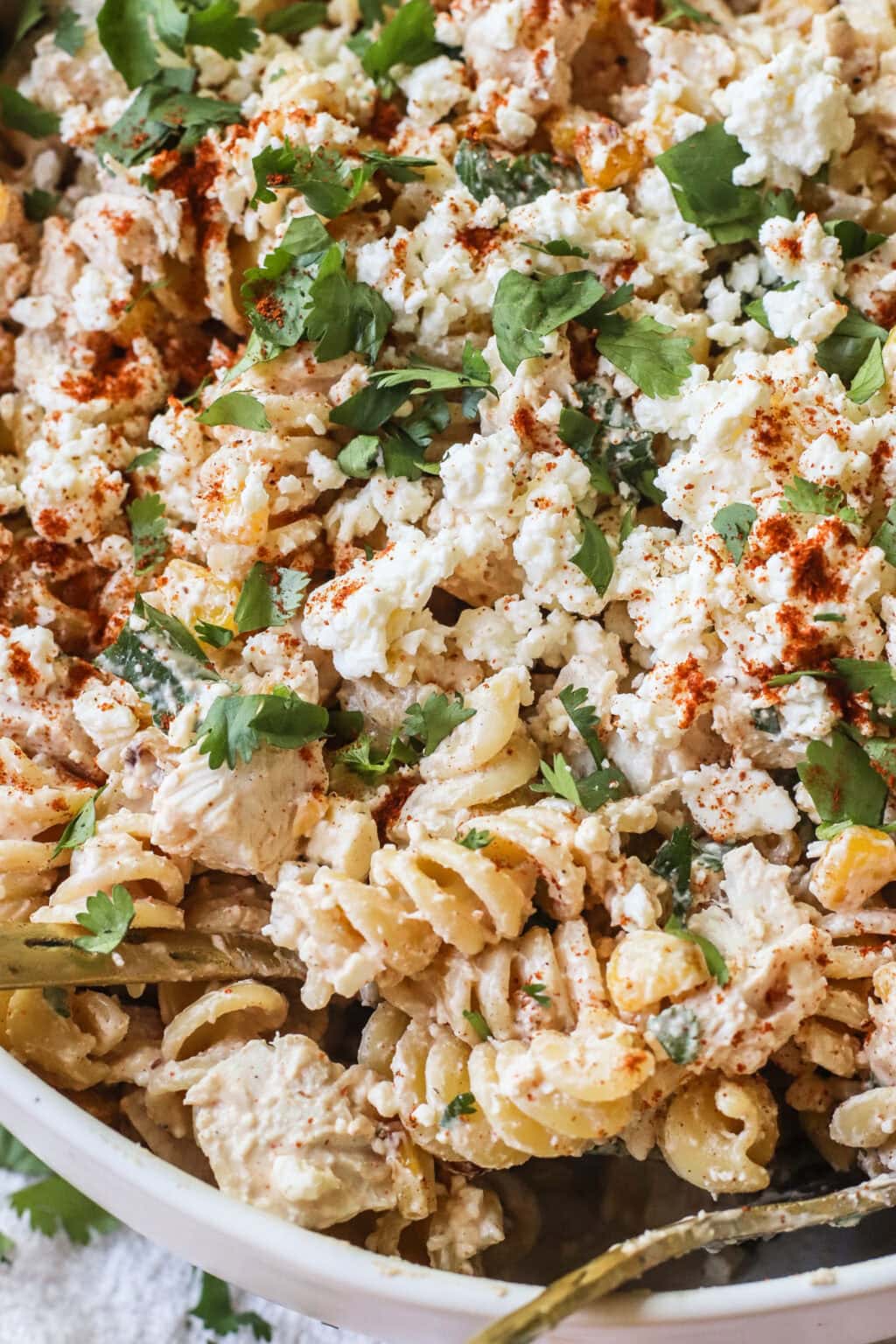 A close-up of a pasta salad made with rotini, shredded chicken, crumbled cheese, chopped cilantro, and sprinkled with paprika. The dish appears creamy and is served in a white bowl.