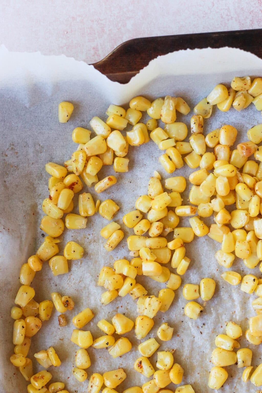 Golden roasted corn kernels scattered on a parchment-lined baking tray, lightly browned and seasoned, with some empty space and a corner of the tray visible.