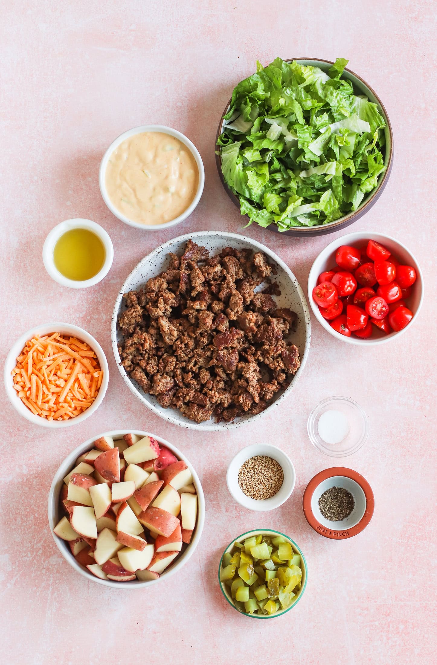 Overhead view of bowls containing ground beef, chopped lettuce, halved cherry tomatoes, diced pickles, shredded cheddar cheese, cubed red potatoes, a creamy dressing, olive oil, sesame seeds, and salt, all on a pink surface.