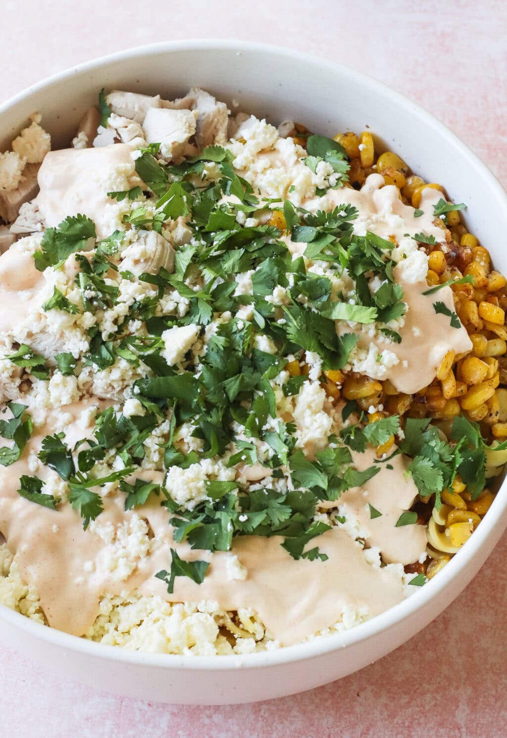A bowl filled with corn, pieces of chicken, crumbled cheese, creamy sauce, and chopped cilantro, arranged on top of noodles.