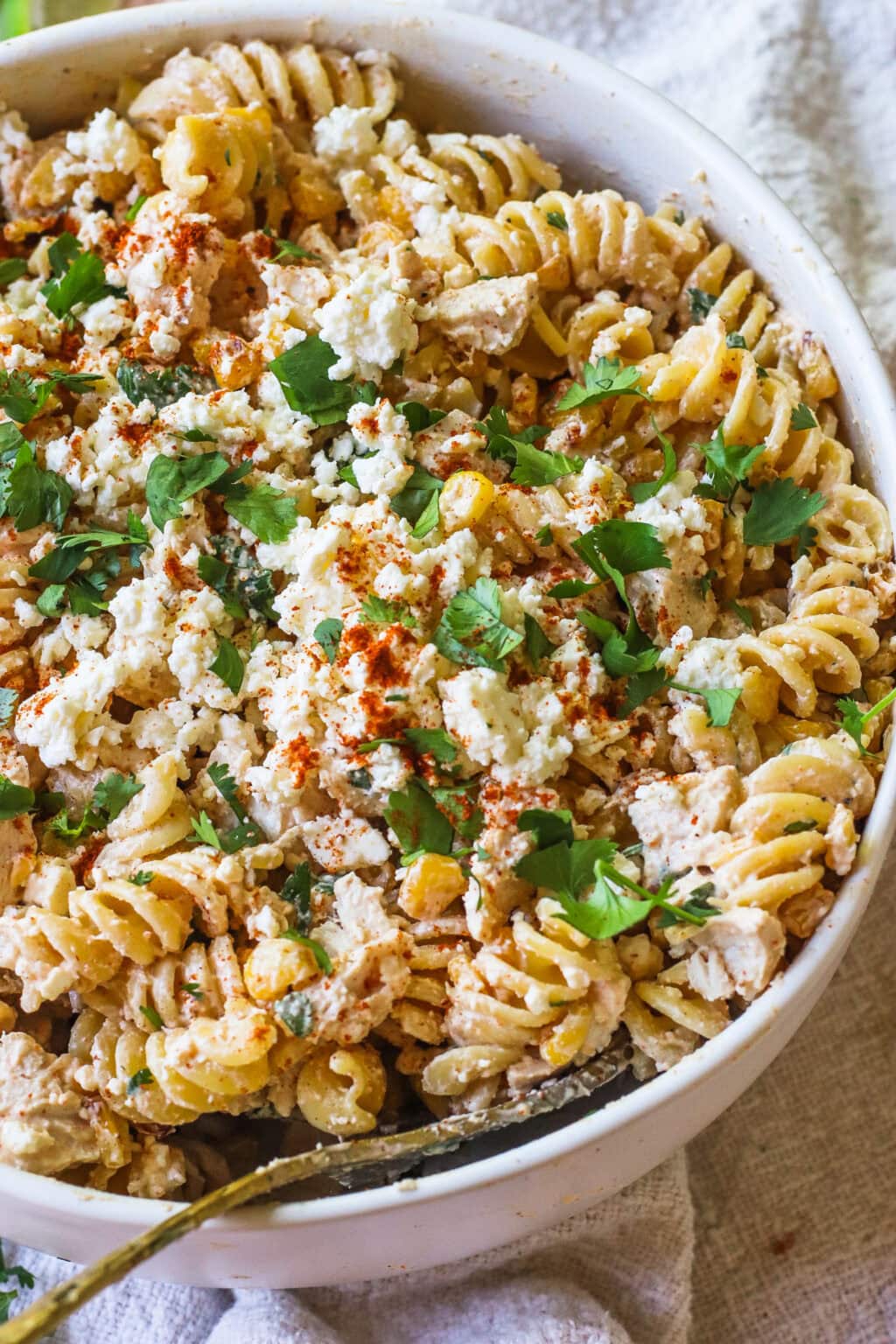 A bowl of rotini pasta mixed with crumbled cheese, corn, fresh cilantro, and sprinkled with paprika, on a light-colored cloth.
