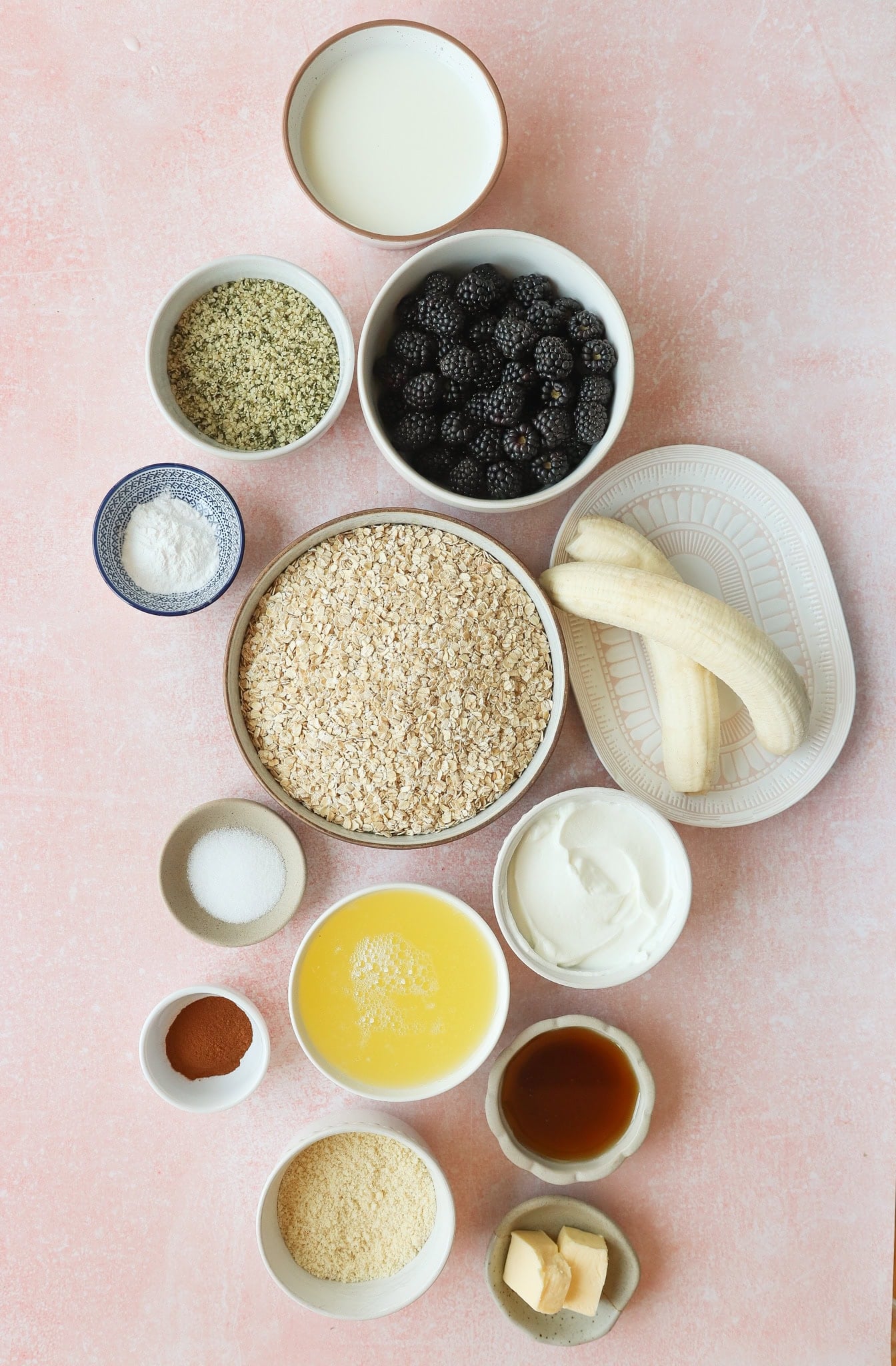 A flat lay of bowls and plates with ingredients including rolled oats, blackberries, banana, greek yogurt, milk, melted butter, hemp seeds, cinnamon, vanilla, baking powder, maple syrup, sugar, and a pinch of salt on a pink surface.