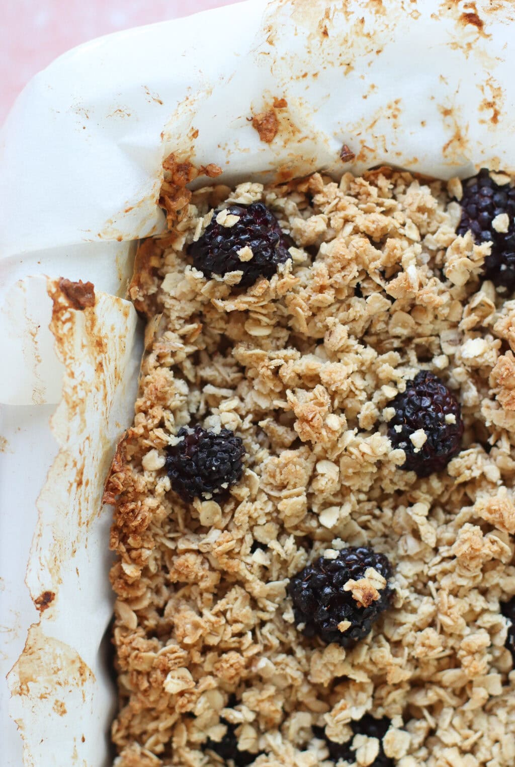 A close-up of a baked blackberry oat crumble in a parchment-lined baking dish, showing golden oats and whole blackberries on top.