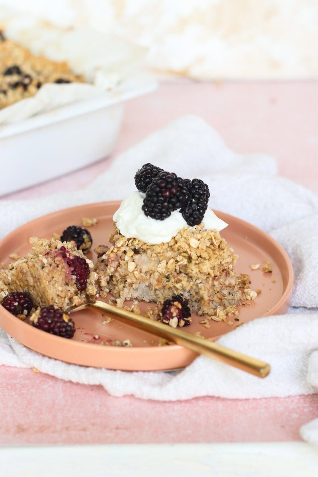 A slice of oat crumble topped with whipped cream and fresh blackberries sits on a pink plate with a gold fork. More blackberries and oat crumble crumbs are scattered around. A baking dish is visible in the background.
