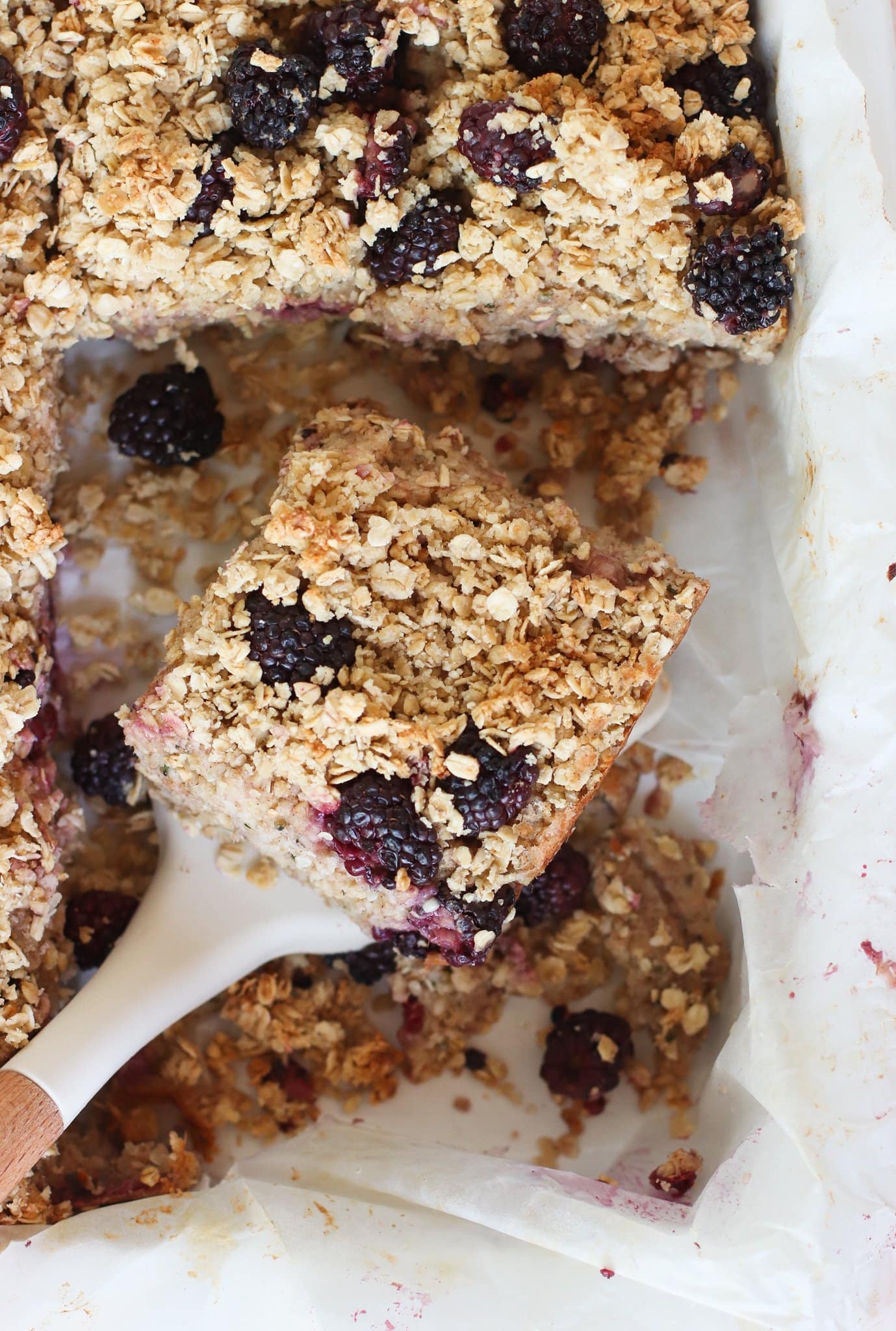 A close-up of a baked oat bar with blackberries being lifted from a parchment-lined pan. Crumbled oats and berries are visible on top and around the bar.