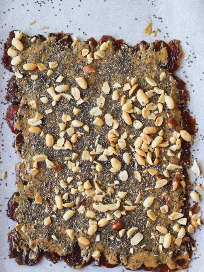 A rectangular sheet of parchment paper topped with a layer of dates, spread with peanut butter, and sprinkled with chia seeds and chopped peanuts.