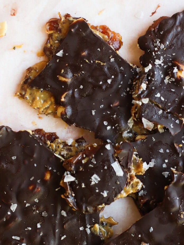 Chocolate-covered brittle pieces topped with flaky sea salt, revealing layers of nuts and a caramel-like filling on a light surface.