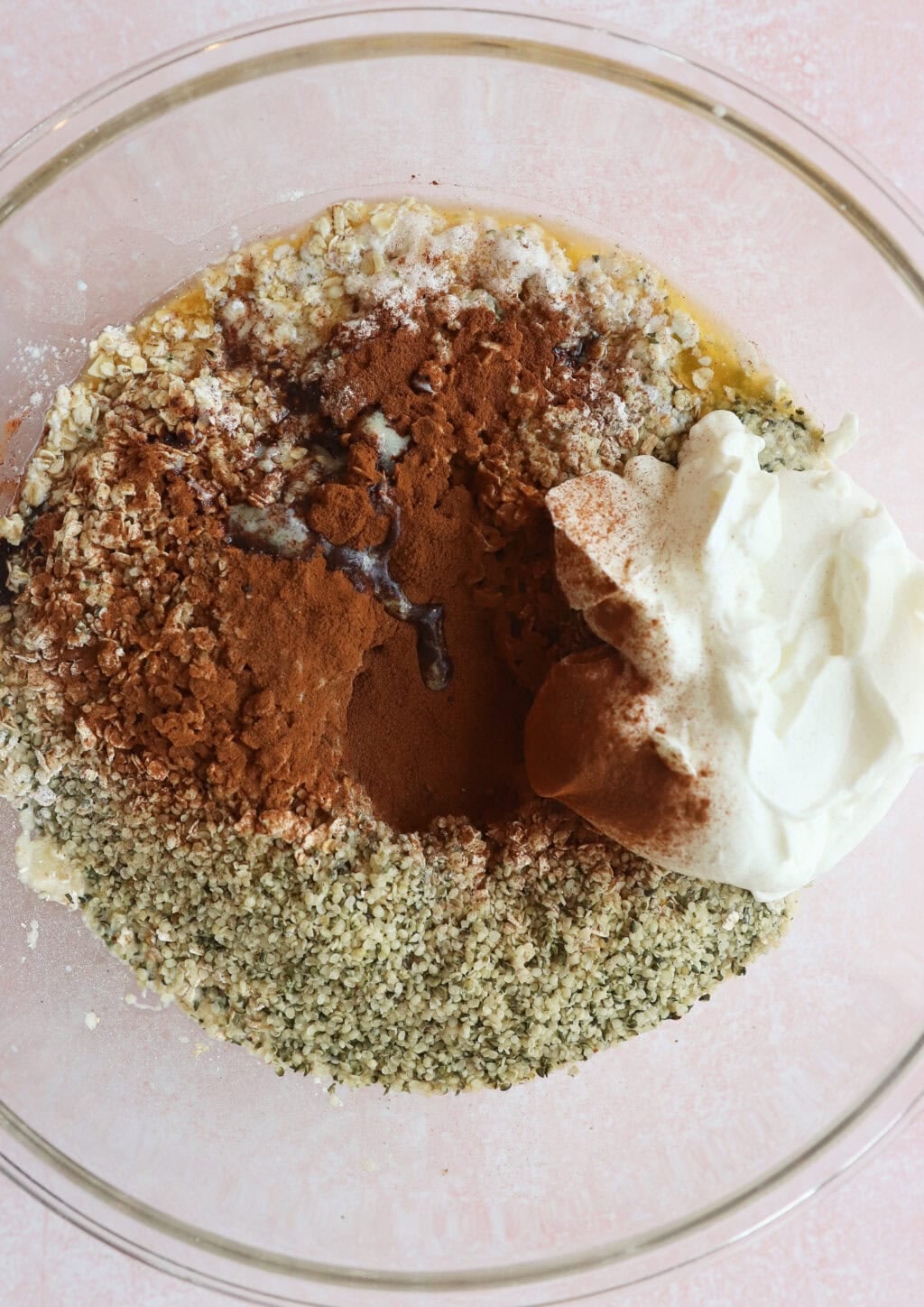 A glass bowl containing oats, ground cinnamon, hemp seeds, Greek yogurt, and a small amount of dark liquid on a light pink surface.