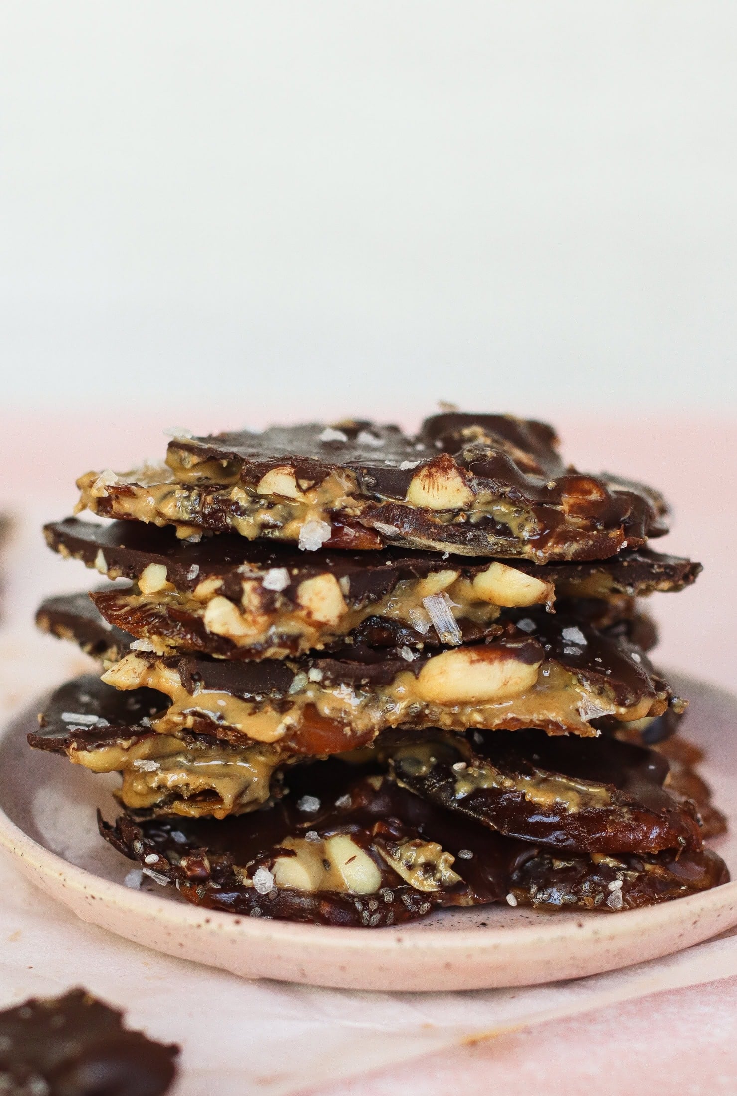 A stack of chocolate-covered nut brittle pieces on a pink plate, sprinkled with flaky sea salt. The brittle shows layers of nuts and dates, with a glossy chocolate coating.