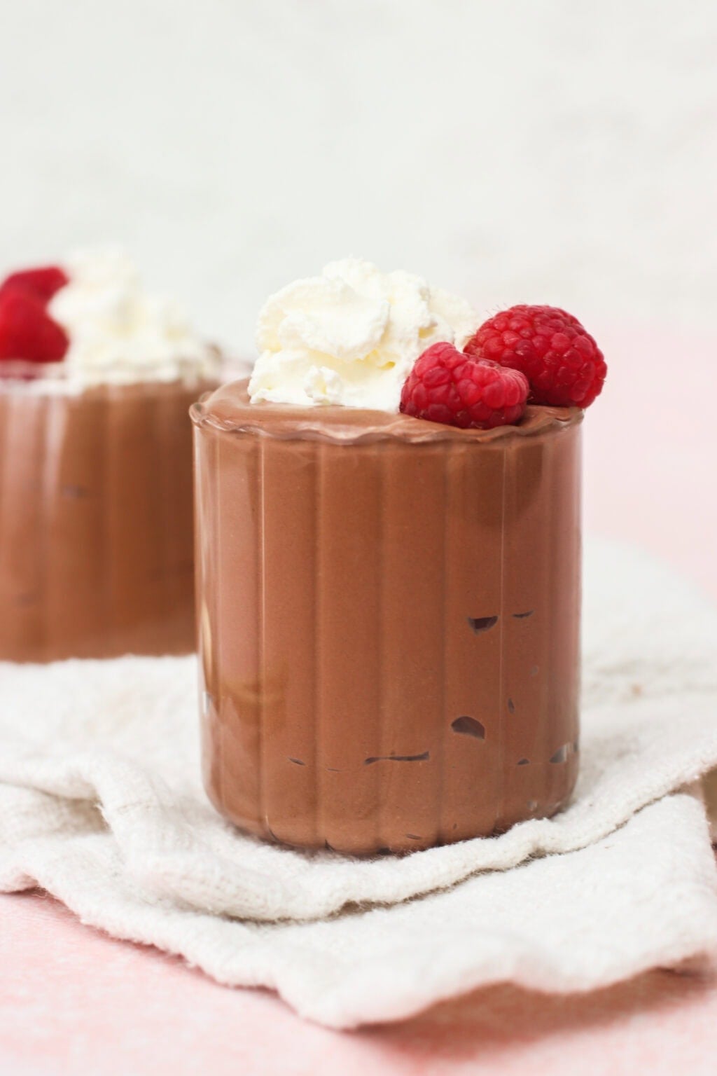 A glass of chocolate mousse topped with whipped cream and fresh raspberries, placed on a light textured cloth, with another similar dessert in the blurred background.