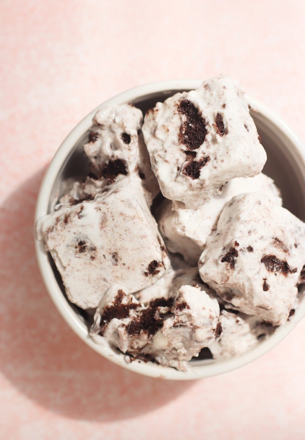 A white bowl filled with square pieces of cookies and cream marshmallows, featuring visible cookie chunks, placed on a light pink textured surface.