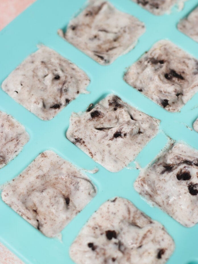 A close-up of a blue silicone mold filled with a cookies and cream mixture, featuring visible chocolate cookie pieces and creamy white base in each square compartment.