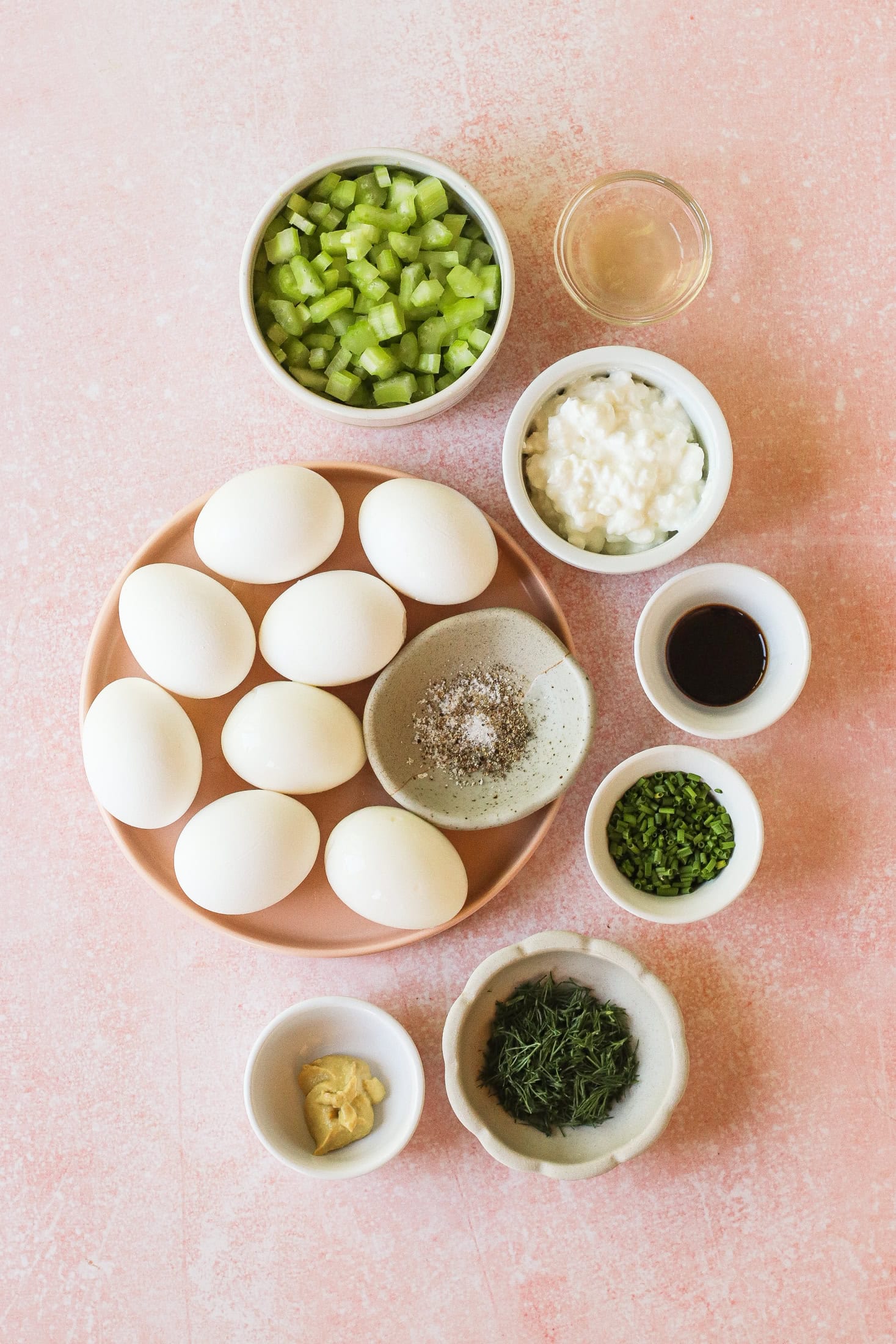 A flat lay of ingredients on a pink surface: hard-boiled eggs, chopped celery, cottage cheese, chives, dill, vinegar, mustard, Worcestershire sauce, and a small dish of salt and pepper.