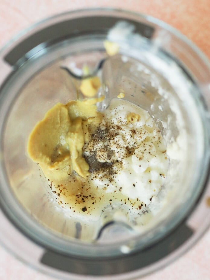 A blender with cottage cheese, mustard, black pepper, and seasonings inside, viewed from above on a pink countertop.