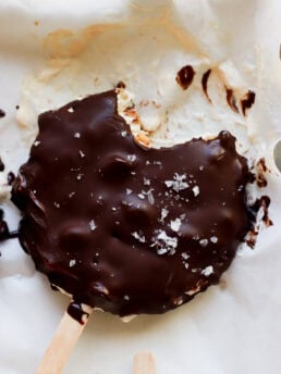 A chocolate-covered ice cream bar with a bite taken out, sprinkled with flaky sea salt, rests on parchment paper with some melted chocolate and ice cream visible around it.