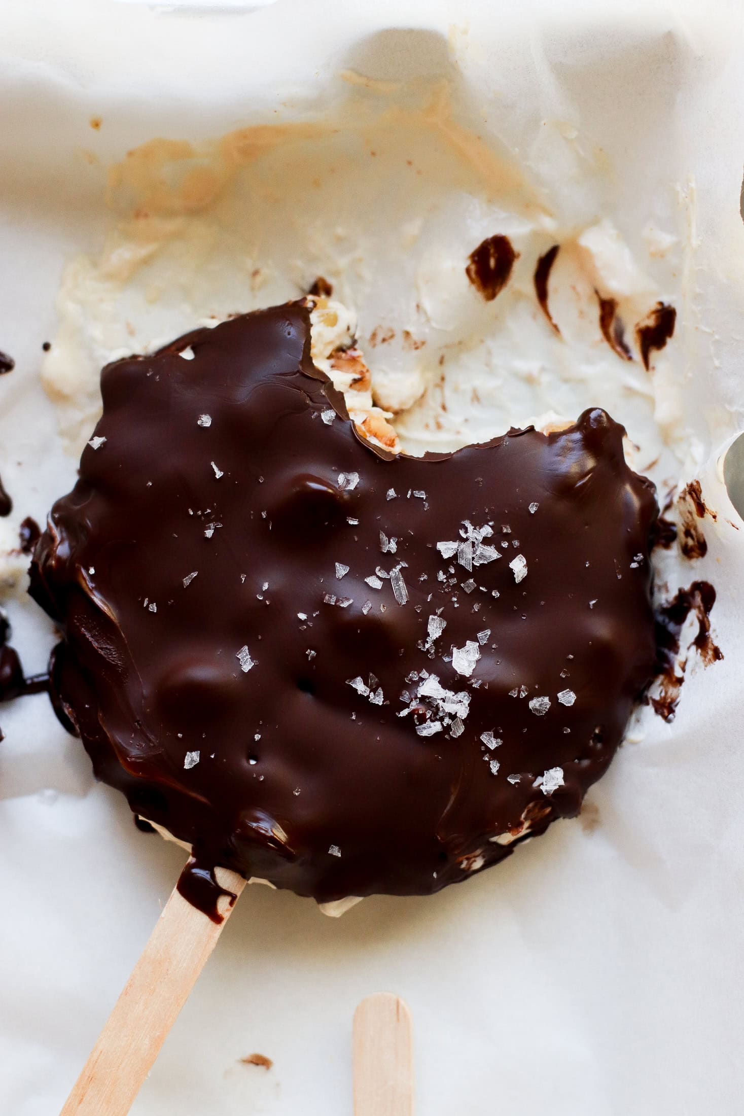 A chocolate-covered ice cream bar with a bite taken out, sprinkled with flaky sea salt, rests on parchment paper with some melted chocolate and ice cream visible around it.