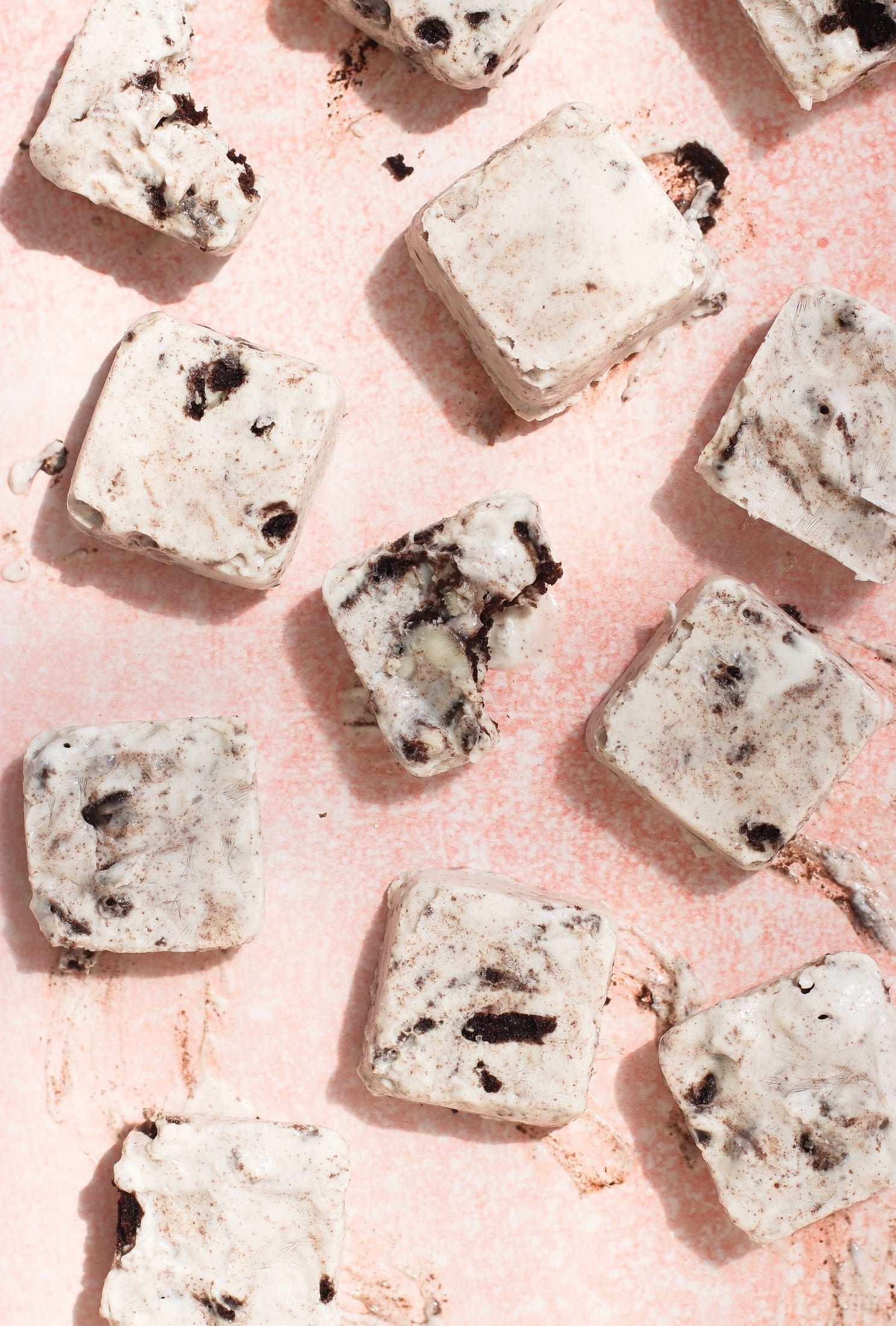 Squares of cookies and cream greek yogurt scattered on a light pink surface, with visible chocolate cookie chunks in the creamy white ice cream. One square has a bite taken out of it.