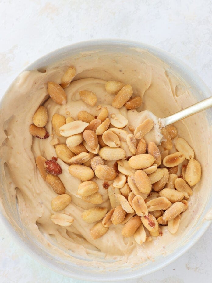 A bowl of creamy peanut butter mixture with roasted peanuts on top, being stirred with a spoon.