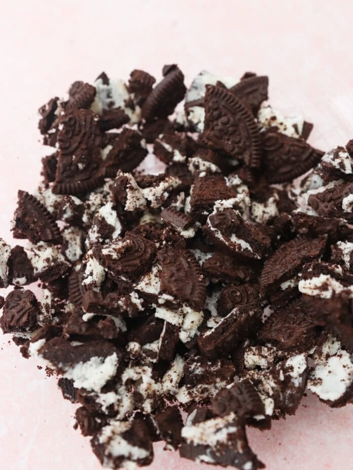 A pile of crushed chocolate sandwich cookies with white cream filling scattered on a light pink surface.
