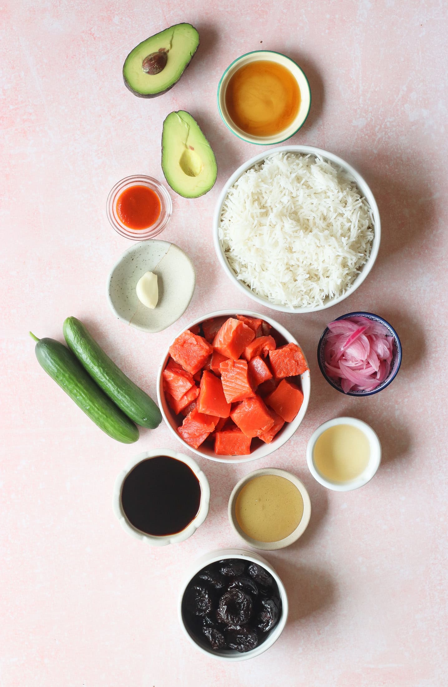 Flat lay of ingredients on a pink surface, including white rice, cubed salmon, cucumbers, avocado halves, soy sauce, prunes, garlic, pickled onions, hot sauce, and small bowls of various sauces.