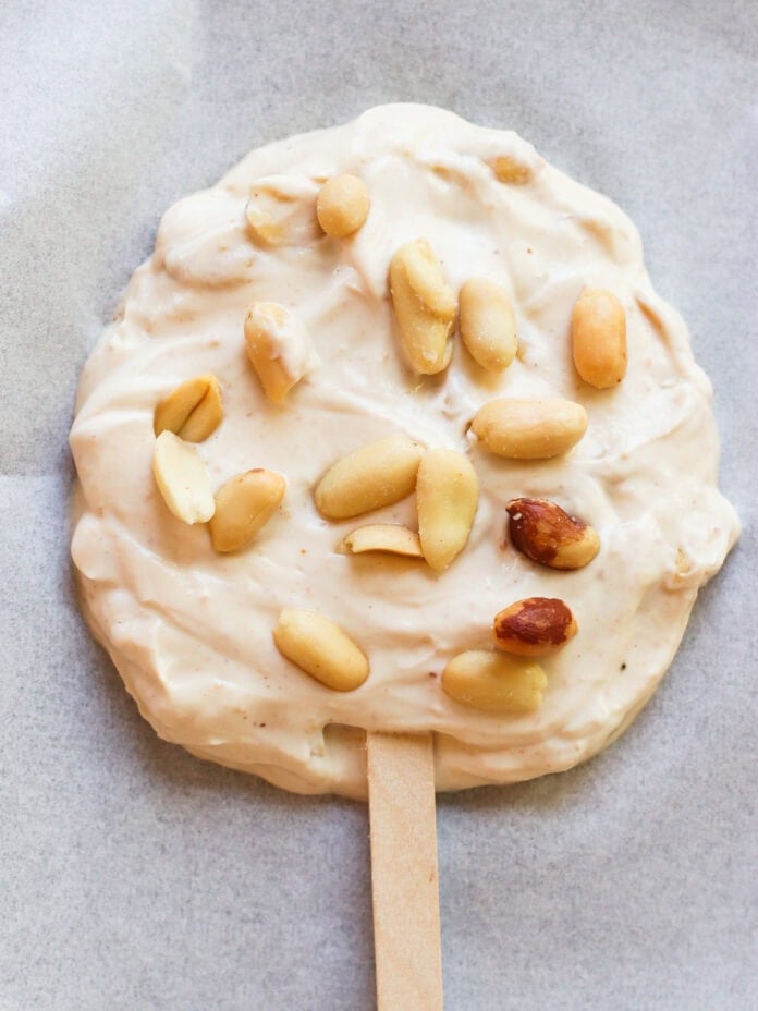A round, creamy frozen treat on a wooden stick, topped with whole peanuts, sits on a sheet of parchment paper.