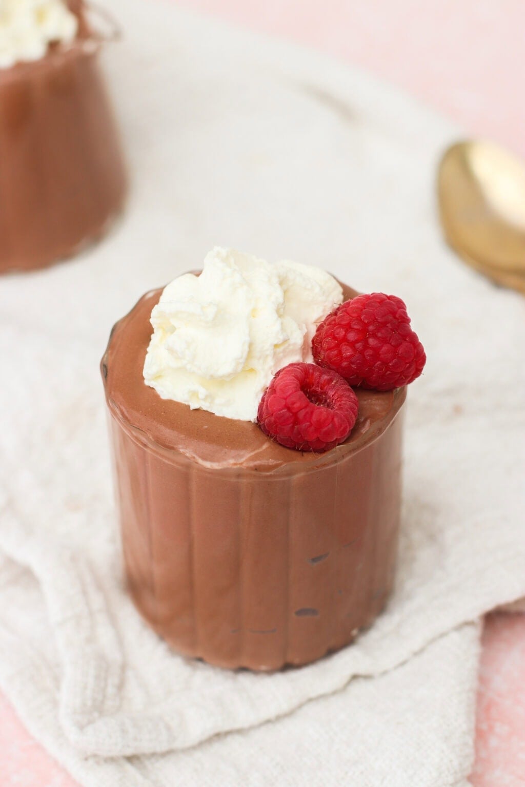 A glass of chocolate mousse topped with whipped cream and two fresh raspberries, placed on a light cloth napkin with a gold spoon in the background.