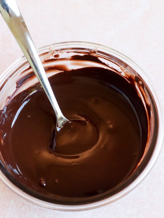 A metal spoon rests in a glass bowl filled with smooth, melted chocolate on a light pink surface.